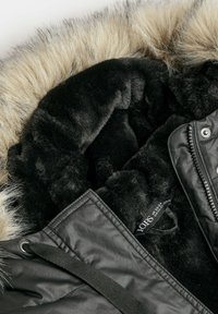 Svart parkas med fuskpäls, som har en mjuk, svart fleecefoder och en tygetikett. Yttermaterialet har en slät, matt yta.