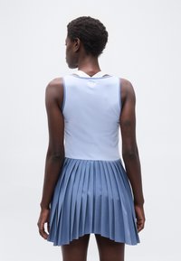 Γυναίκα με κοντά σγουρά μαλλιά φοράει αμάνικη ανοιχτό μπλε μπλούζα και πλισέ σκουρόχρωμη μπλε φούστα, στέκεται με την πλάτη προς τα εμπρός σε λευκό φόντο.