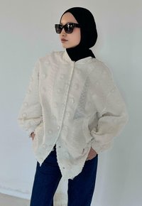 Veste blanche texturée avec motifs en relief à pois, fermeture à boutons et manches froncées, associée à un jean foncé et des lunettes de soleil.