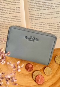 Portafoglio in pelle blu chiaro con zip, impresso con "Gusti Leder studio", posizionato su una superficie testurizzata con monete e un libro sullo sfondo.
