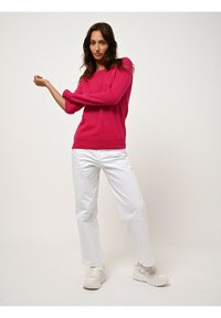 Maglione fucsia lavorato a maglia con maniche ampie, abbinato a jeans a gamba dritta bianchi e sneakers chiare, su uno sfondo uniforme.