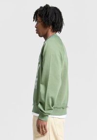 Felpa verde con finitura texturizzata, caratterizzata da un collo girocollo e maniche raglan, orlo sfrangiato e logo laterale bianco. Abbinata a pantaloni beige.