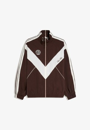 Veste de survêtement zippée marron et blanche avec un motif chevron blanc, logo sur la poitrine gauche, et inscription "Stay Late" sur la poitrine droite.