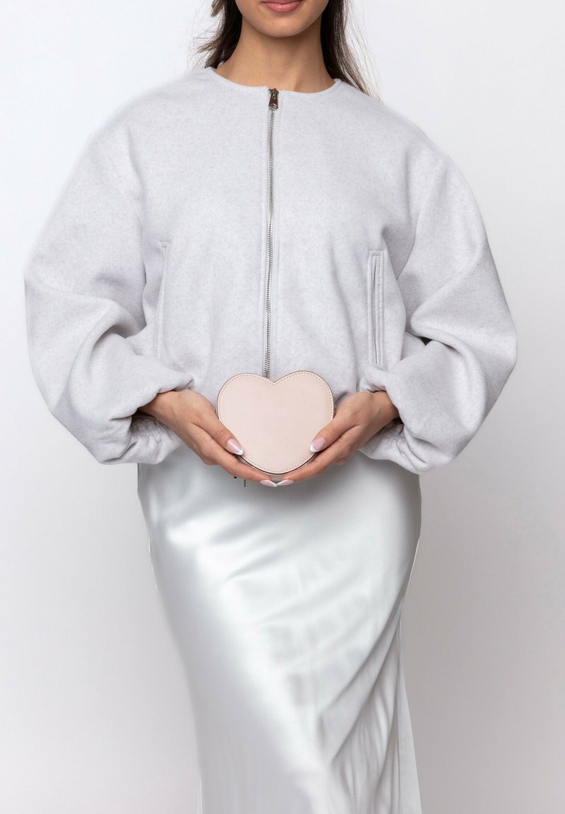 Gray fleece bomber jacket, oversized fit, zippered front. Holding a pink heart-shaped leather bag with stitched edges. White satin skirt.