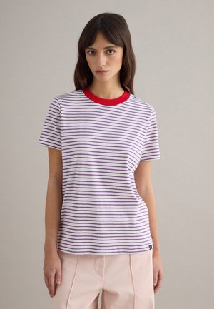 Mujer joven con camiseta de rayas blancas y moradas con cuello rojo y pantalones rosa claro, de pie contra un fondo claro y liso.