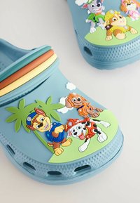 Sabots en caoutchouc bleu avec une bande rayée multicolore et des motifs d'animaux en relief. Présente un palmier et des chiens de dessin animé sur une base verte.