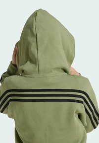 Sudadera verde hecha de una tela suave, con una gran capucha y tres franjas horizontales negras en la parte posterior. Textura suave y corte casual.