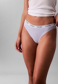 Tanga en coton violet clair avec une ceinture élastique noire portant un logo. Texture lisse et couvrance minimale, conçu pour le confort et l'ajustement.