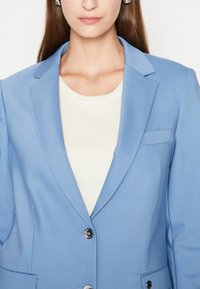 Světle modrý blazer se strukturovaným designem, jednoduchým knoflíkovým zapínáním a malou kapsou na levé straně, vrstvený přes krémový top.