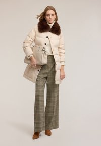 Kobieta ubrana w beżowy pikowany płaszcz z ciemnym futrzanym kołnierzem, szerokie spodnie w kratę, brązowe botki na obcasie i trzymająca beżową torebkę.
