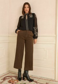 Melnā caurspīdīgā blūze ar ziedu izšuvumiem, atslēgas caurumu detaļa, kopā ar brūnām platām biksēm. Melnas papēžu zābaki piešķir kontrastu.