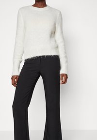 Person som bär en vit fluffig långärmad tröja och svarta högmidjade vida byxor mot en enfärgad vit bakgrund.