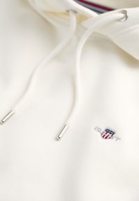 White hoodie made of cotton, featuring a kangaroo pocket, drawstrings with silver tips, and a small embroidered logo. Clean design.