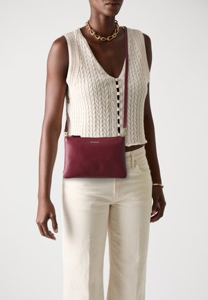 Red leather crossbody bag with a textured finish, gold zipper, and strap, held by a person wearing a knit top and light pants.