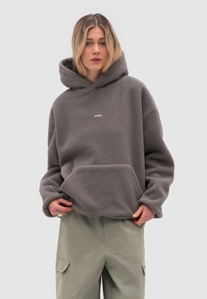 Kvinde iført oversized grå fleece-hoodie med forlomme og lysegrå cargobukser, stående mod en ensfarvet lys baggrund.