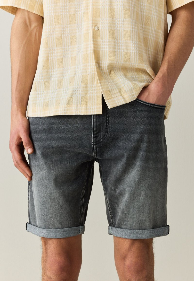 Homme portant un short en denim noir délavé retroussé et une chemise à manches courtes jaune à carreaux avec une main dans la poche.