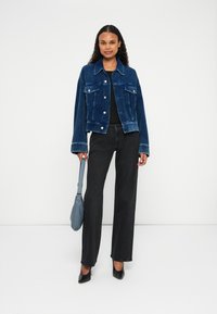 Giubbotto di velluto a coste blu con tasche frontali, chiusura a bottoni e cuciture a contrasto; abbinato a jeans neri a gamba larga e tacchi neri.