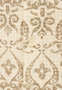 Κοντινό πλάνο μπεζ υφάσματος με καφέ περίτεχνα λουλουδένια και σπειροειδή σχέδια και ένα στρογγυλό μπεζ κουμπί με τέσσερις τρύπες ραμμένο πάνω στο ύφασμα.