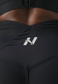 Czarne legginsy sportowe z wysokim stanem o gładkiej fakturze. Zauważalna biała logo z tyłu, widoczne szwy.