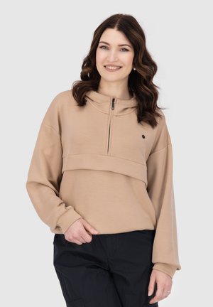 Donna con capelli castani ondulati, indossa un felpa beige con mezza zip e pantaloni neri, sorridente su uno sfondo chiaro e uniforme.