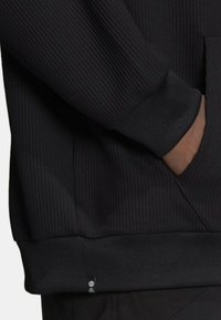 Svart sweatshirt med ribbat mönster, åtsittande ärmslut och en framficka. Har en liten logotag vid fållen. Materialet verkar mjukt och hållbart.