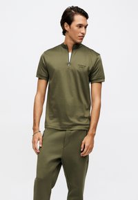 Άνδρας που φοράει ελαιοπράσινο μπλουζάκι με κοντά μανίκια και φερμουάρ μπροστά και αντίστοιχο παντελόνι, κοιτάζοντας προς τα αριστερά του, μπροστά σε απλό λευκό φόντο.