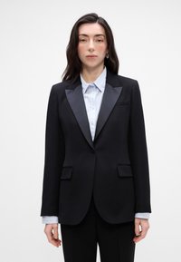 Zwart tuxedo-blazer met satijnen revers, twee voorzakken en gestructureerde schouders, gedragen over een lichtblauw gestreept shirt.
