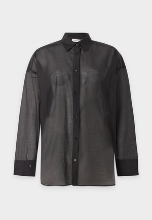 Person iført løs, gennemskinnelig sort skjorte med knapper og matchende gennemskinnelige bukser med elastiske manchetter, stående og vendt mod fronten på ensfarvet baggrund.