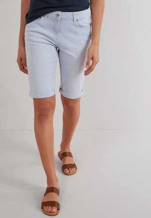 Femme portant un short bleu clair retroussé et des sandales plates marron à double lanière, marchant sur un sol uni.