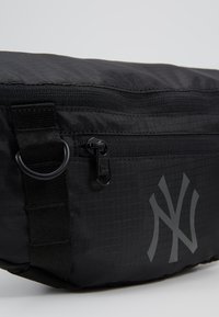 Sac banane en nylon noir avec une texture lisse, doté d'une poche zippée, d'un matériel en métal et d'une impression de logo gris bien en vue.