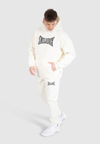 Krämfärgad hoodie med svart "SMILODOX"-text, tillsammans med matchande sweatpants. Tyget verkar mjukt, med en avslappnad passform och framfickor.