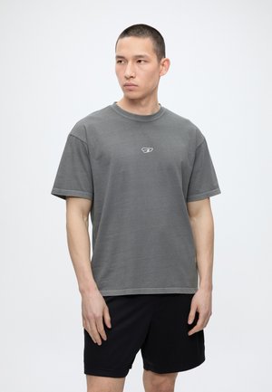 Ung mand med kort hår iført en grå t-shirt og sorte shorts, kigger til siden mod en ensfarvet hvid baggrund.