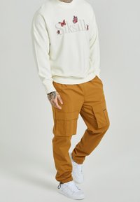 Vit sweatshirt med broderade fjärilar och logotyp, tillsammans med bruna cargo-byxor med stora sidofickor och vita sneakers.