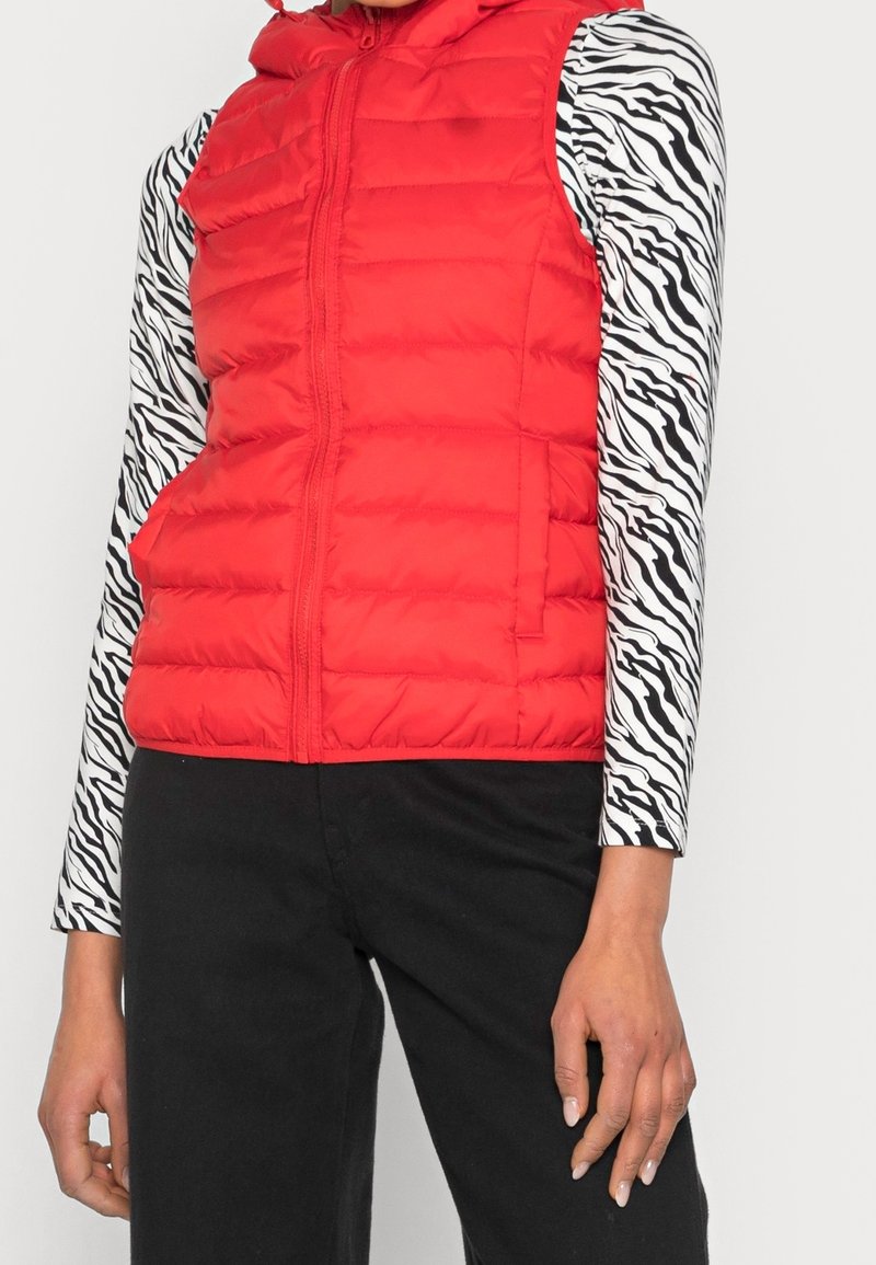 Rode gewatteerde vest met een ritssluiting aan de voorkant, voorzien van een capuchon; gedragen over een zwart-wit gestreepte top met lange mouwen en een zwarte broek.