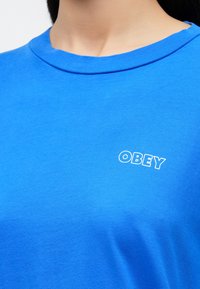 Μπλε βαμβακερό μπλουζάκι με στρογγυλή λαιμόκοψη και κοντά μανίκια. Διακοσμημένο με λευκό λογότυπο "OBEY" στο στήθος. Ομαλή υφή.