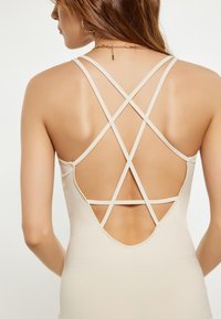 Beige mouwloze body met een crisscross rugontwerp met dunne bandjes en een diepe uitsnijding. Soepele textuur en een aansluitende silhoutte.