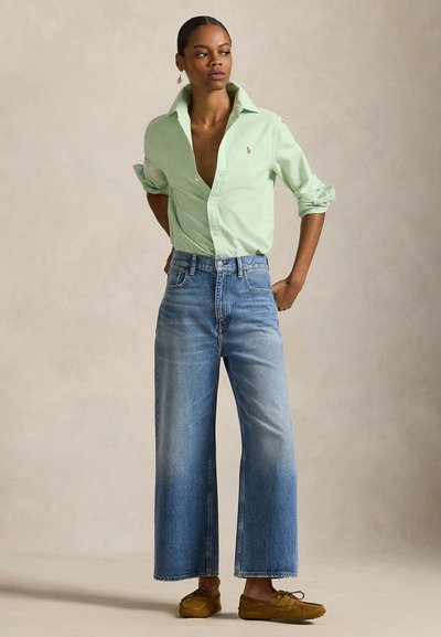 Vrouw draagt een lichtgroene blouse met knopen en opgerolde mouwen, hoge, brede blauwe spijkerbroek en bruine mocassinschoenen, staand met handen achter.