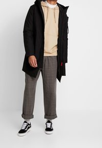 Czarna parka, pod spodem beżowa bluza z kapturem, spodnie w kratę oraz czarne sneakersy. Elementy charakterystyczne to kaptur, zamek błyskawiczny oraz czerwone akcenty.