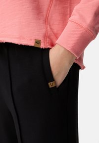 Hellrosa Sweatshirt mit strukturierter Stoffoberfläche, rohem Saum und gerippten Bündchen. Schwarze Hose mit Seitentaschen und braunen Markenetiketten.