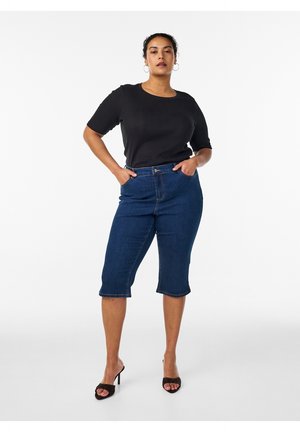 Kvinde stående med hænderne i lommerne iført sort kortærmet top, mørkeblå knælange denimshorts og sorte åbne hæle på hvid baggrund.