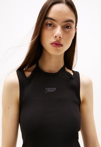 Black ribbed tank top with cut-out shoulder details, featuring a gray "tommy jeans" logo on the chest. Soft, form-fitting fabric.