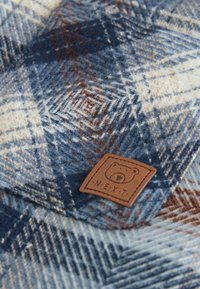 Tissu à carreaux bleu, blanc et marron avec une texture douce. Présente une étiquette en cuir tan avec un logo d'ours et " .NEXT. " brodé dessus.