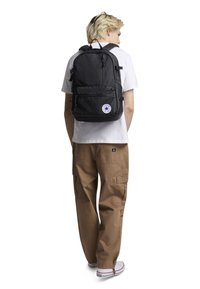 Svart ryggsekk med teksturert materiale, flere glidelåsrom, polstrede stropper og et rundt logo-patch på forsiden.