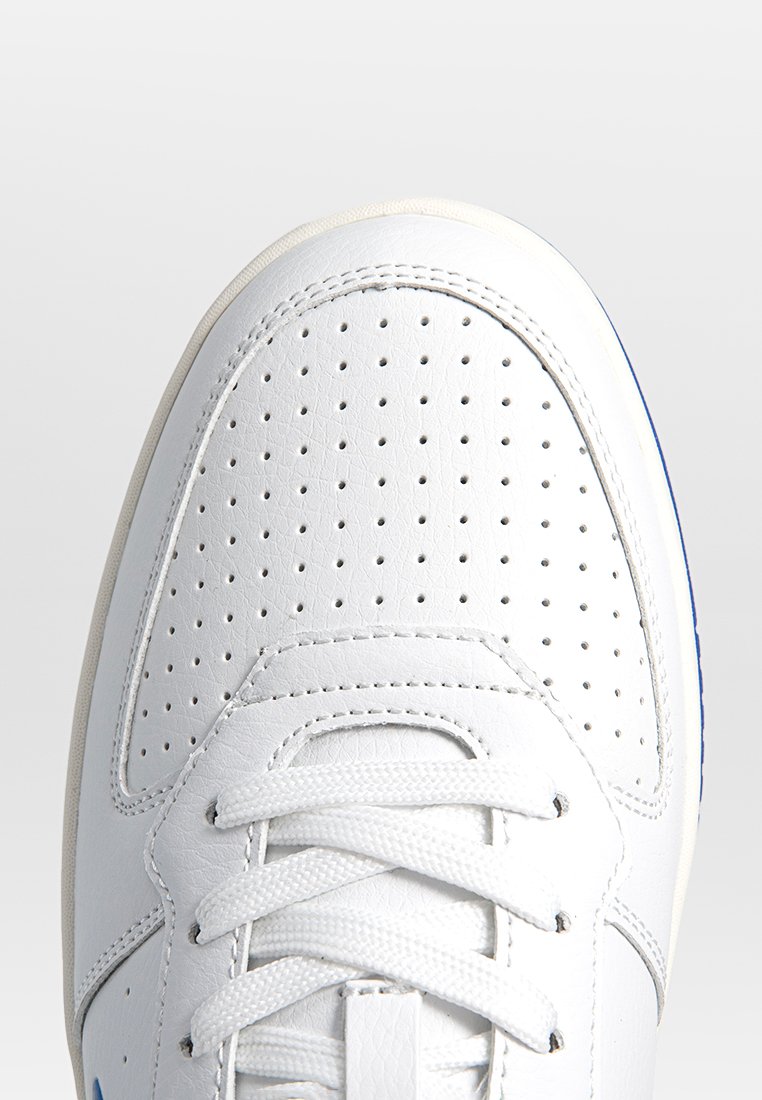 Zapatilla de cuero blanca con puntera perforada, cordones blancos y detalles cosidos. El lateral tiene un acento azul cerca de la suela.
