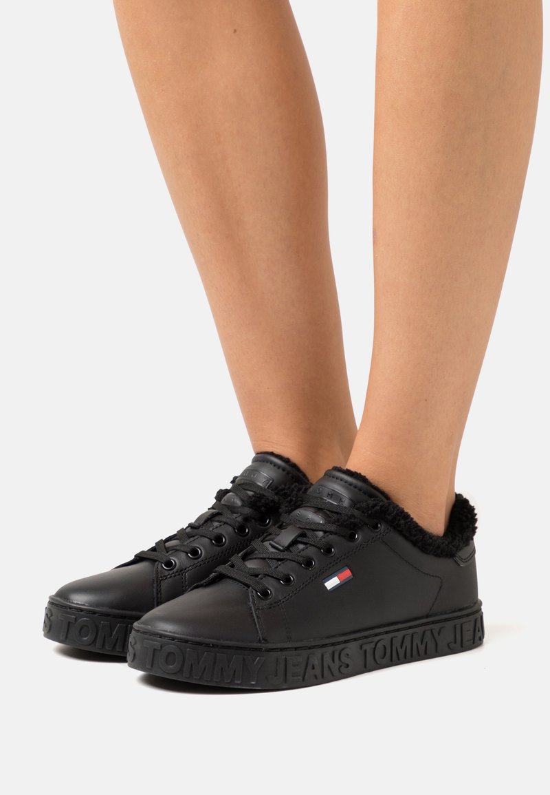 Baskets basses noires avec lacets, doublure en fourrure douce et logo embossé "Tommy Jeans" sur la semelle, portées pieds nus.