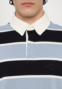 Polo skjorte med horisontale blå, hvite og marineblå striper. Har en ribbet hvit krage og broderte logo-detaljer i marineblå.
