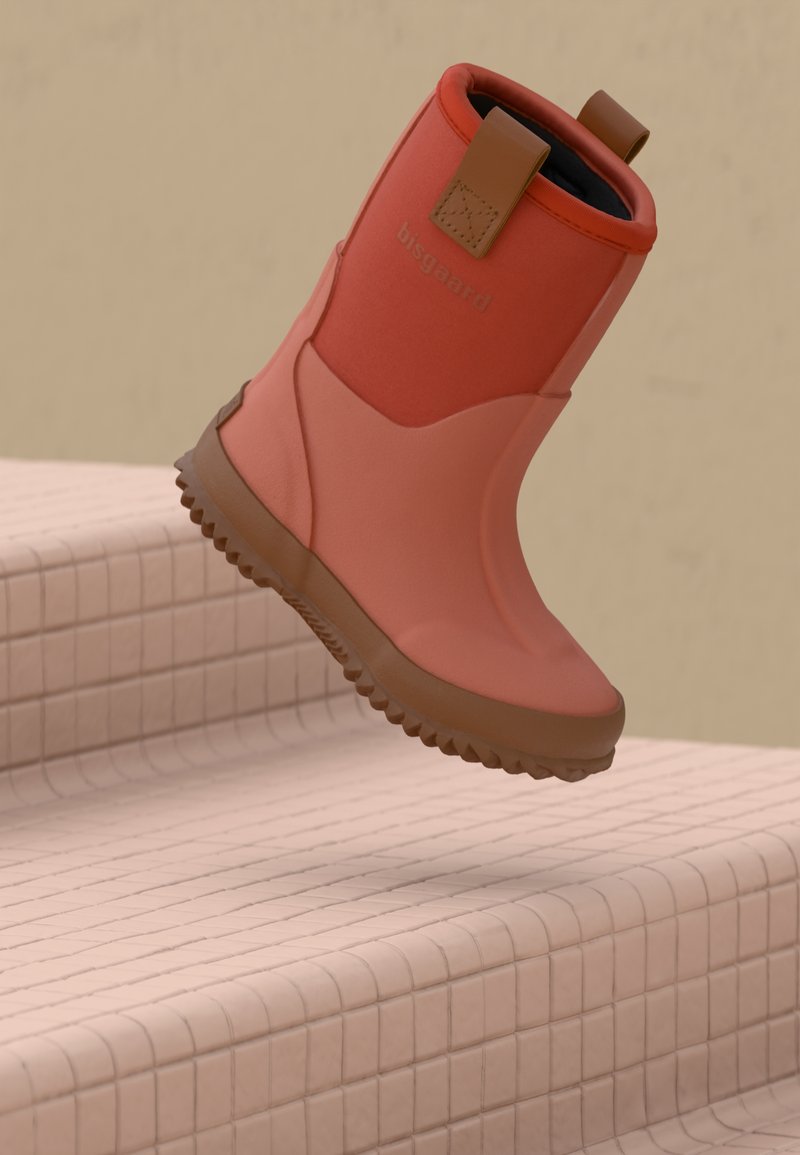 Botte en caoutchouc rouge avec semelle marron et poignées de tirage, marquée "bisgaard", suspendue au-dessus de marches carrelées beiges.