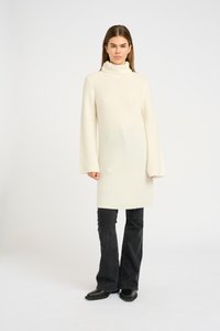 Abito a collo alto in maglia color crema, con maniche ampie, che si ferma poco sopra il ginocchio. Abbinato a jeans neri a zampa d'elefante e stivaletti.