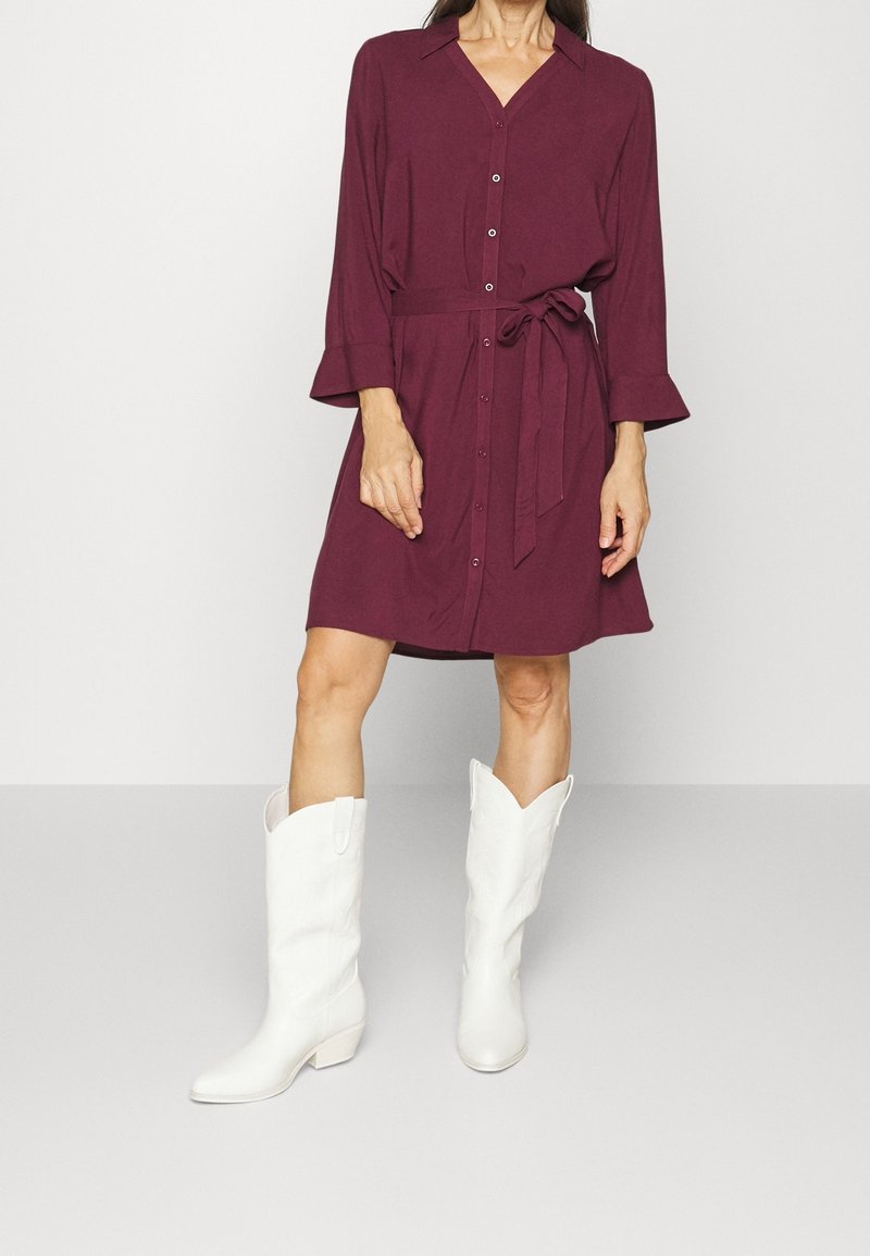 Femme portant une robe boutonnée bordeaux longueur genou avec une ceinture à la taille et des bottes blanches hautes jusqu'aux genoux, debout devant un fond uni.