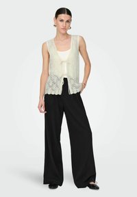 Lace-gehaakt mouwloos crèmekleurig vest met een striksluiting aan de voorkant, gecombineerd met losse zwarte broeken en platte zwarte schoenen. Eenvoudig ontwerp en lichte textuur.